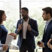 Confident African male leader standing telling diverse colleagues about new project, boss lead briefing for different age workgroup team employees in office. Sharing information and leadership concept