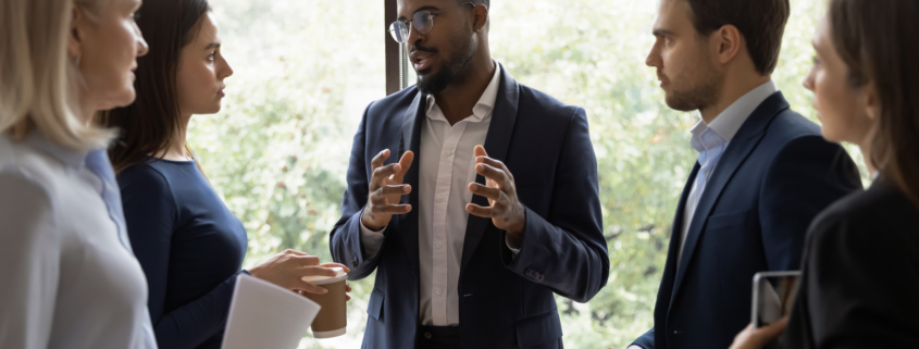 Confident African male leader standing telling diverse colleagues about new project, boss lead briefing for different age workgroup team employees in office. Sharing information and leadership concept