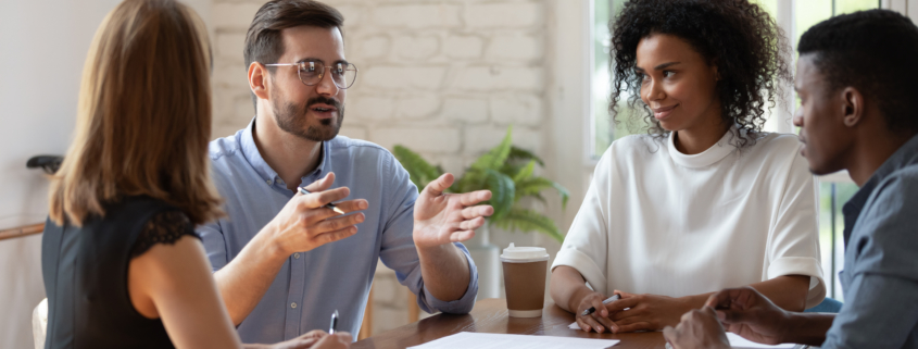 Diverse business partners brainstorming, discussing project at meeting, sitting at table in boardroom, colleagues listening to businessman sharing ideas, confident coach mentor training staff